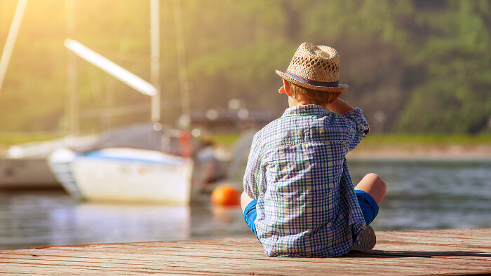 Ein kleiner Bub sitzt auf einem Holzsteg und blickt auf ein Segelboot