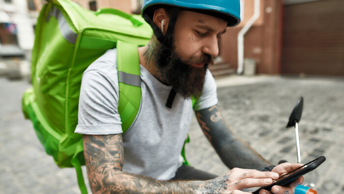 Essenslieferant mit Helm und gro&szlig;em gr&uuml;nen Thermo-Rucksack sucht auf seinem Handy, sitzt dabei auf einem (E-)Moped