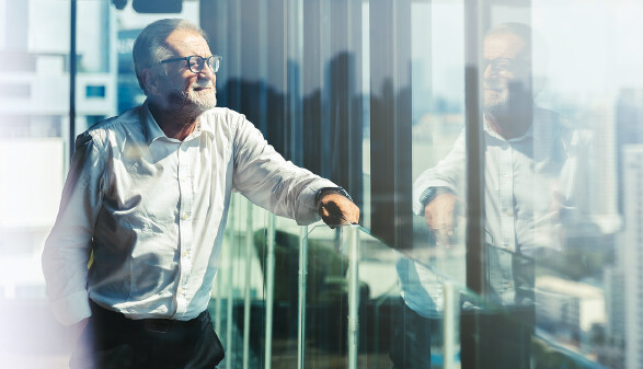 Ein &auml;lterer Gesch&auml;ftsmann steht in einer modernen Lounge hoch oben in einem B&uuml;roturm und blickt nach rechts.