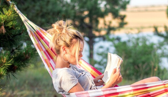 Junge Frau liegt in einer H&auml;ngematte im Gr&uuml;nen am See oder Fluss und liest ein Buch