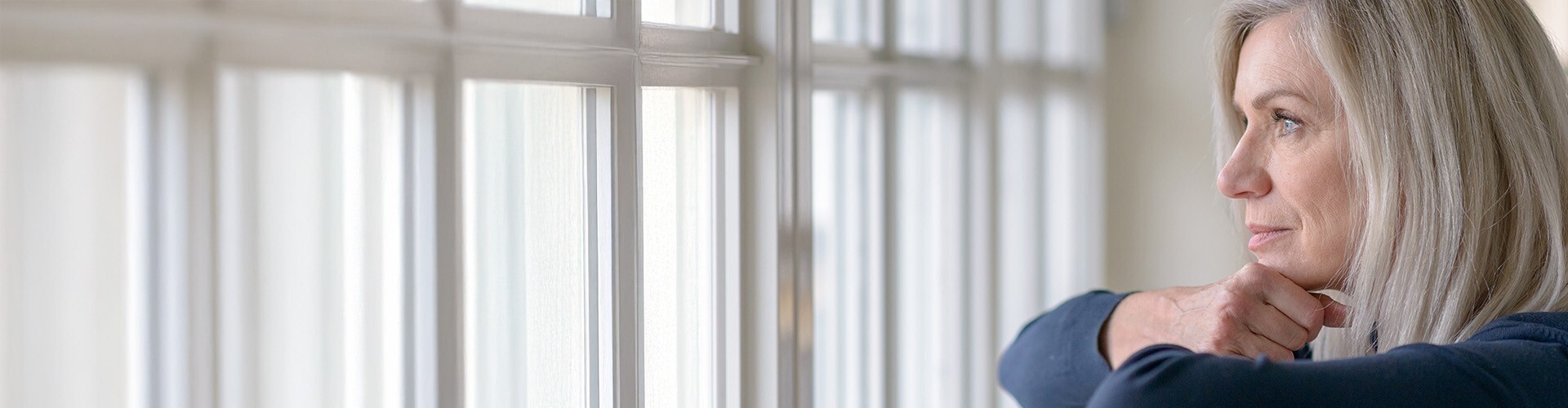 Reife, blonde Frau blickt traurig aus dem Fenster