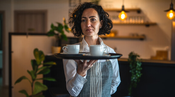 Kellnerin trägt Tablett mit zwei Kaffeetassen mit ernstem Blick durch ein Café