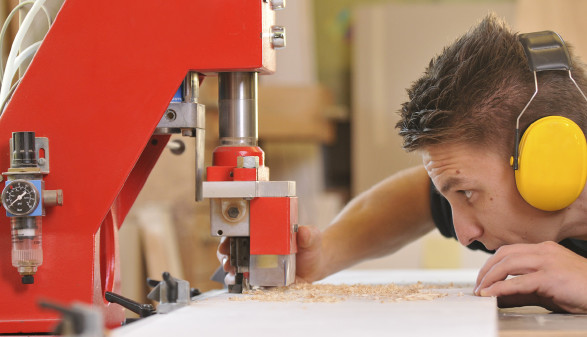 Ein männlicher Jugendlicher mit Gehörschutz steht an einer Werkbank und bearbeitet ein Holzbrett mit einer Fräsmaschine.