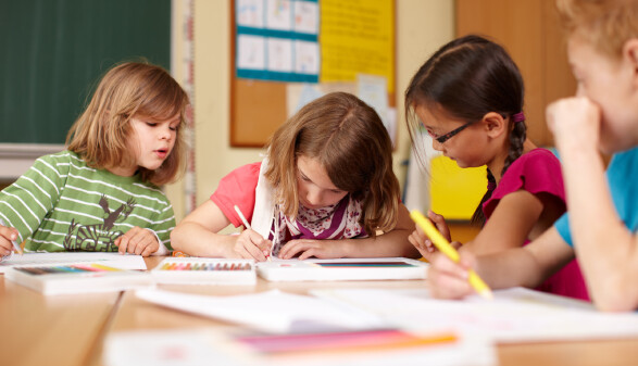 Vier Volksschulkinder sitzen nebeneinander an einer Schulbank und malen konzentriert