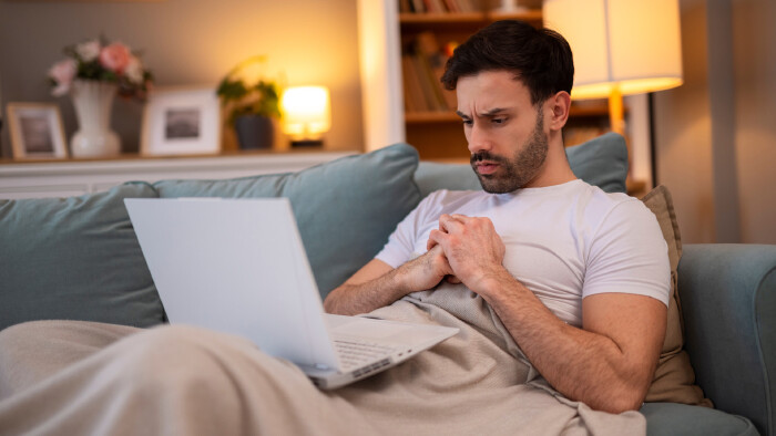 Mann, zugedeckt, halb liegend auf einer Couch mit Laptop auf dem Scho&szlig;, schaut verzweifelt. 